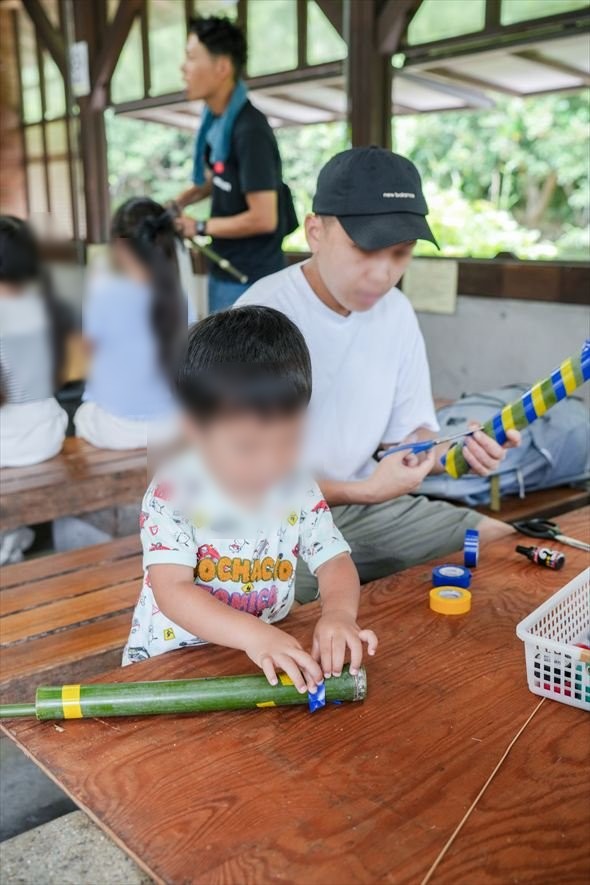 屋根の下のテーブルで、親子で竹の水鉄砲を組み立てる